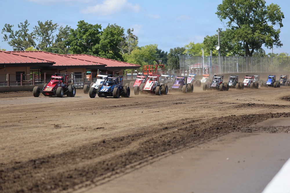 A large field of open-wheel sprint/midget-style dirt track race cars are lined up and racing on a dirt oval track. The cars are spread across the width of the track, kicking up dust as they accelerate. A red-roofed building and green trees are visible in the background under a partly cloudy sky.