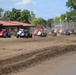 A large field of open-wheel sprint/midget-style dirt track race cars are lined up and racing on a dirt oval track. The cars are spread across the width of the track, kicking up dust as they accelerate. A red-roofed building and green trees are visible in the background under a partly cloudy sky.