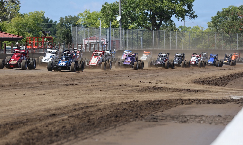 A large field of open-wheel sprint/midget-style race cars lines up on a dirt oval track, appearing to be at the start or restart of a race. The cars are arranged in a tight pack spanning the width of the track, kicking up dust as they accelerate. The venue features a chain-link fence perimeter, grandstand structures, and surrounding trees visible in the background.
