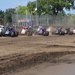 A large field of open-wheel sprint/midget-style race cars lines up on a dirt oval track, appearing to be at the start or restart of a race. The cars are arranged in a tight pack spanning the width of the track, kicking up dust as they accelerate. The venue features a chain-link fence perimeter, grandstand structures, and surrounding trees visible in the background.