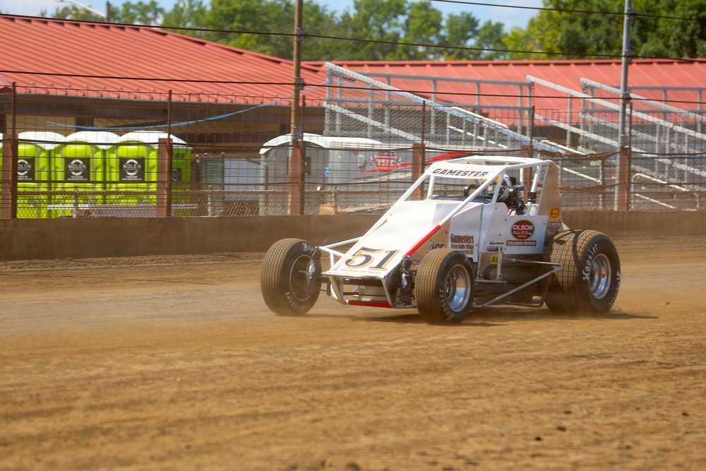 A white open-wheel dirt track racing car numbered 51, sponsored by 'Gamester' and 'Olson', is navigating a dirt oval racing track. The vehicle appears to be a wingless sprint car or midget racer kicking up dust as it moves along the track surface. The setting is a small regional racing facility with grandstands, a red-roofed building, and portable toilets visible in the background.