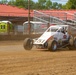 A white open-wheel dirt track racing car numbered 51, sponsored by 'Gamester' and 'Olson', is navigating a dirt oval racing track. The vehicle appears to be a wingless sprint car or midget racer kicking up dust as it moves along the track surface. The setting is a small regional racing facility with grandstands, a red-roofed building, and portable toilets visible in the background.