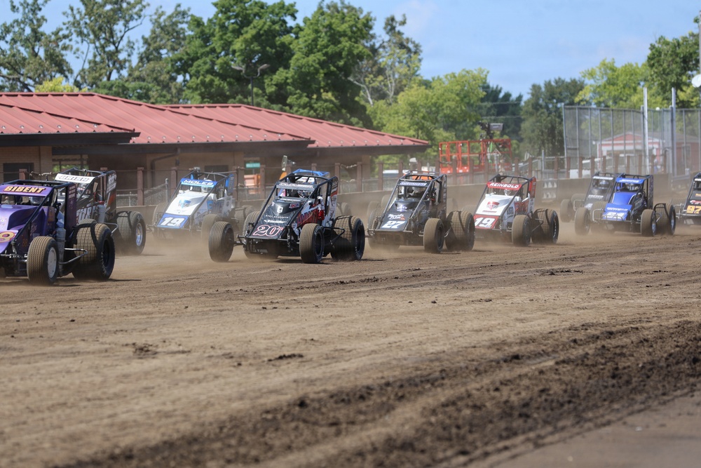 A pack of open-wheel dirt track racing cars (midgets or sprint cars) compete on a dirt oval circuit during what appears to be a race start or early-race grouping. Multiple numbered vehicles are visible kicking up dust on a brown dirt surface, with a red-roofed building and green trees visible in the background. The scene is captured in bright daylight conditions at what appears to be a small regional racing venue.