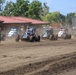 A pack of open-wheel dirt track racing cars (midgets or sprint cars) compete on a dirt oval circuit during what appears to be a race start or early-race grouping. Multiple numbered vehicles are visible kicking up dust on a brown dirt surface, with a red-roofed building and green trees visible in the background. The scene is captured in bright daylight conditions at what appears to be a small regional racing venue.