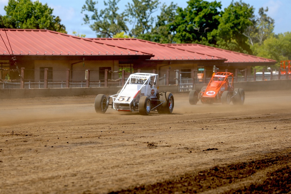 Two open-wheel dirt track racing cars are competing on a dusty dirt oval circuit. The lead car is white and numbered 51 with 'GAMESTER' visible on the roll cage, while an orange car follows closely behind. A red-roofed building and green trees are visible in the background.