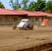 Two open-wheel dirt track racing cars are competing on a dusty dirt oval circuit. The lead car is white and numbered 51 with 'GAMESTER' visible on the roll cage, while an orange car follows closely behind. A red-roofed building and green trees are visible in the background.