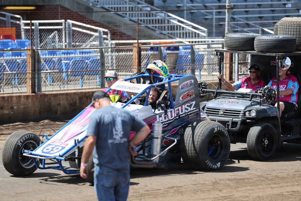 A dirt track racing scene showing a blue and white open-wheel sprint car numbered 69 sponsored by 'Loueusz Automotive Network' and 'Lucas Oil' being pushed or escorted through the pit area. A driver wearing a colorful helmet is seated in the car, while crew members in matching pink and black shirts accompany the vehicle on a John Deere utility cart carrying spare tires. The setting appears to be an indoor or stadium-style dirt track facility with blue grandstand seating visible in the background.