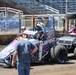 A dirt track racing scene showing a blue and white open-wheel sprint car numbered 69 sponsored by 'Loueusz Automotive Network' and 'Lucas Oil' being pushed or escorted through the pit area. A driver wearing a colorful helmet is seated in the car, while crew members in matching pink and black shirts accompany the vehicle on a John Deere utility cart carrying spare tires. The setting appears to be an indoor or stadium-style dirt track facility with blue grandstand seating visible in the background.