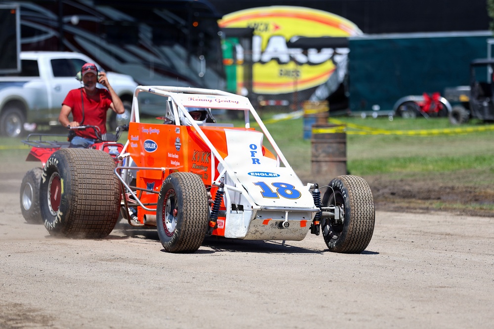 A dirt track racing scene featuring an orange and white open-wheel sprint car numbered 18, sponsored by 'OFR' and 'Engler', driven by Kenny Gentry. The car is being pushed or escorted by a person on an ATV/quad bike on a dirt surface. The setting appears to be a racing pit or staging area at a motorsport event.