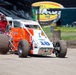 A dirt track racing scene featuring an orange and white open-wheel sprint car numbered 18, sponsored by 'OFR' and 'Engler', driven by Kenny Gentry. The car is being pushed or escorted by a person on an ATV/quad bike on a dirt surface. The setting appears to be a racing pit or staging area at a motorsport event.