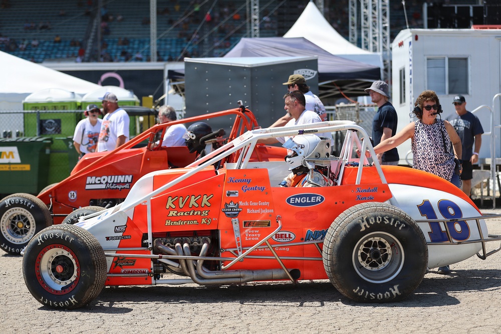 A pit lane or staging area at a racing event featuring two orange open-wheel sprint/champ cars. The foreground car is number 18, sponsored by KVKK Racing from Henderson, KY, with driver Kenny Gentry seated in the cockpit wearing a helmet. Several crew members, officials, and bystanders are visible around the vehicles.