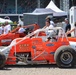 A pit lane or staging area at a racing event featuring two orange open-wheel sprint/champ cars. The foreground car is number 18, sponsored by KVKK Racing from Henderson, KY, with driver Kenny Gentry seated in the cockpit wearing a helmet. Several crew members, officials, and bystanders are visible around the vehicles.