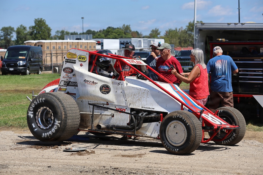 A dirt track racing scene showing a white and red open-wheel sprint car (midget car) with Hoosier tires and various sponsor decals, parked in a pit area. Several people are gathered around the vehicle, including a helmeted driver seated in the car and crew/family members conversing nearby. The setting appears to be a rural racing facility on a sunny day.