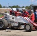 A dirt track racing scene showing a white and red open-wheel sprint car (midget car) with Hoosier tires and various sponsor decals, parked in a pit area. Several people are gathered around the vehicle, including a helmeted driver seated in the car and crew/family members conversing nearby. The setting appears to be a rural racing facility on a sunny day.