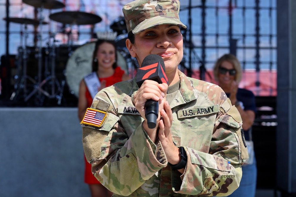 A U.S. Army female soldier with the last name 'ALVA' (partially visible) is holding a microphone and speaking or performing at what appears to be a public event. She is wearing Army Combat Uniform (ACU) in MultiCam pattern with a Staff Sergeant rank insignia on her cap and a U.S. flag patch on her left sleeve. In the background, a drum kit, a woman wearing a pageant sash and crown, and another woman in casual attire are visible.