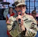 A U.S. Army female soldier with the last name 'ALVA' (partially visible) is holding a microphone and speaking or performing at what appears to be a public event. She is wearing Army Combat Uniform (ACU) in MultiCam pattern with a Staff Sergeant rank insignia on her cap and a U.S. flag patch on her left sleeve. In the background, a drum kit, a woman wearing a pageant sash and crown, and another woman in casual attire are visible.