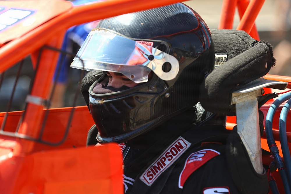 A racing driver is seated inside an open-wheel or dirt track race car, wearing full safety gear. The driver is equipped with a black Simpson-branded helmet and racing suit, surrounded by the orange roll cage structure of the vehicle. The image appears to be taken in a pit or staging area prior to or during a race event.
