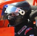 A racing driver is seated inside an open-wheel or dirt track race car, wearing full safety gear. The driver is equipped with a black Simpson-branded helmet and racing suit, surrounded by the orange roll cage structure of the vehicle. The image appears to be taken in a pit or staging area prior to or during a race event.