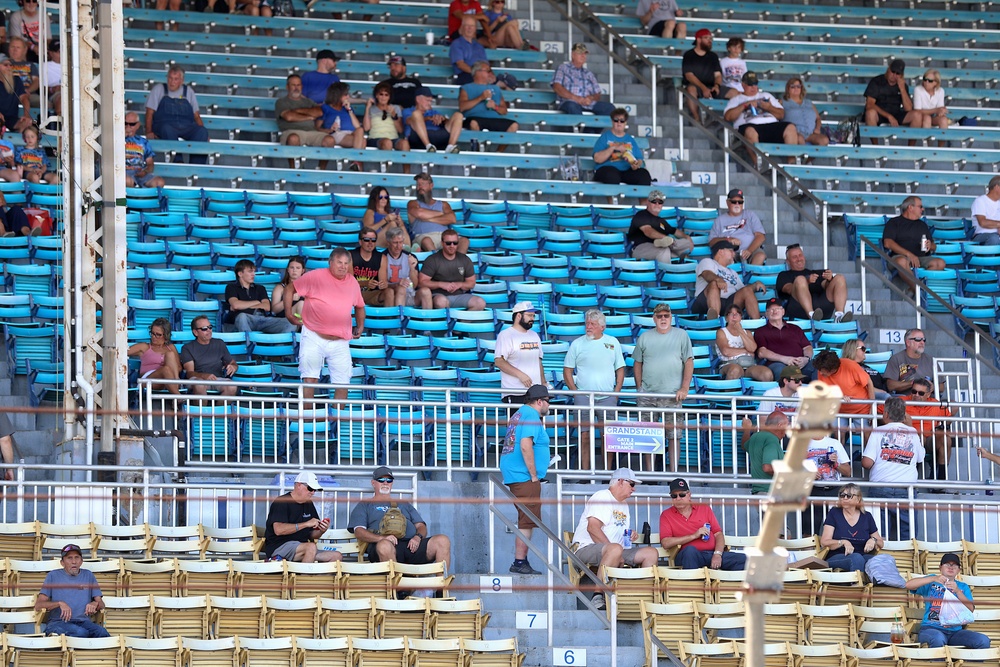 A partially filled grandstand at what appears to be a motorsports racing venue, with spectators spread across multiple seating sections. The stands feature two distinct seating areas: light blue plastic seats in the upper sections and tan/beige folding chairs in the lower sections. Attendance is sparse with many empty seats visible throughout the venue.