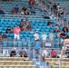 A partially filled grandstand at what appears to be a motorsports racing venue, with spectators spread across multiple seating sections. The stands feature two distinct seating areas: light blue plastic seats in the upper sections and tan/beige folding chairs in the lower sections. Attendance is sparse with many empty seats visible throughout the venue.