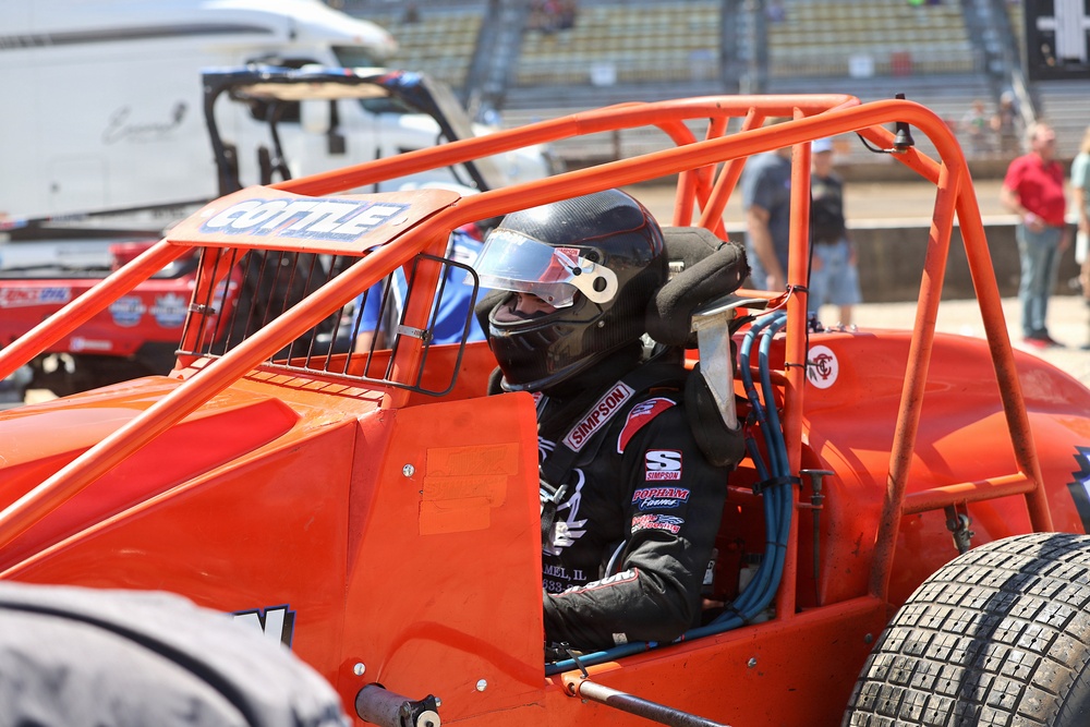 A racing driver sits in an open-wheel sprint car or midget race car with a bright orange roll cage frame. The driver is wearing a black Simpson racing helmet and black racing suit with visible sponsor logos. The scene appears to be in a pit or staging area at a racing venue with grandstands visible in the background.