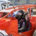 A racing driver sits in an open-wheel sprint car or midget race car with a bright orange roll cage frame. The driver is wearing a black Simpson racing helmet and black racing suit with visible sponsor logos. The scene appears to be in a pit or staging area at a racing venue with grandstands visible in the background.