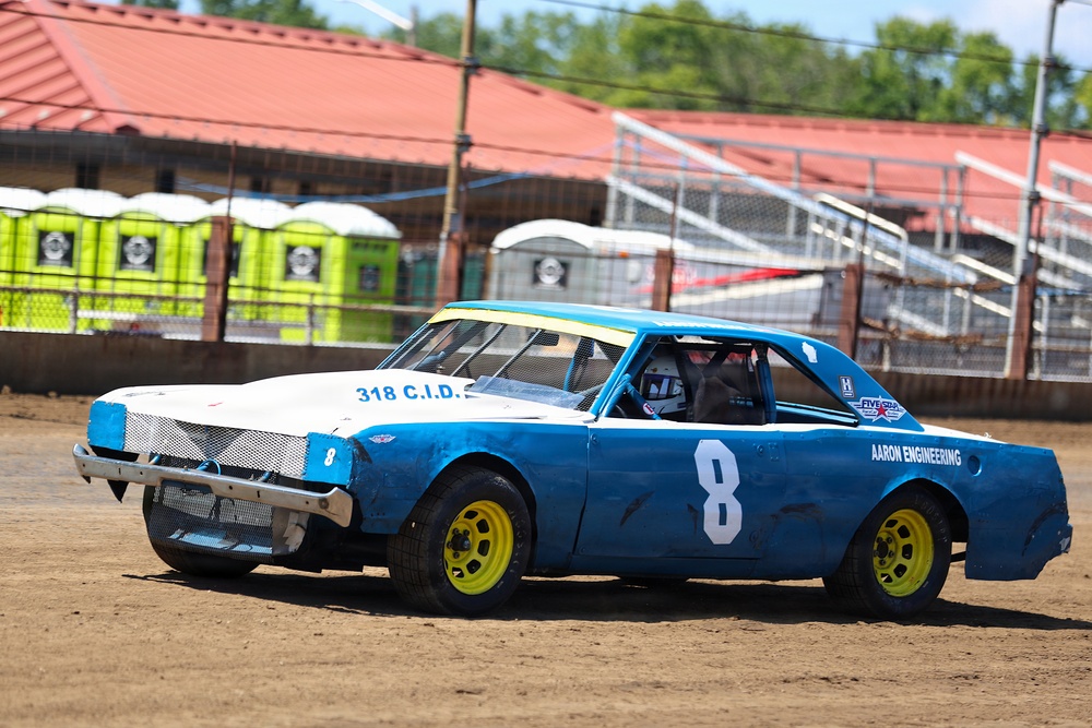 A blue and white dirt track race car numbered '8' is competing on a dirt oval racing surface. The car displays sponsor markings including 'Aaron Engineering' and '318 C.I.D.' on its body, with yellow painted wheels. The background shows portable toilets, grandstand bleachers, and a red-roofed structure typical of a small regional racing venue.