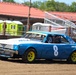 A blue and white dirt track race car numbered '8' is competing on a dirt oval racing surface. The car displays sponsor markings including 'Aaron Engineering' and '318 C.I.D.' on its body, with yellow painted wheels. The background shows portable toilets, grandstand bleachers, and a red-roofed structure typical of a small regional racing venue.