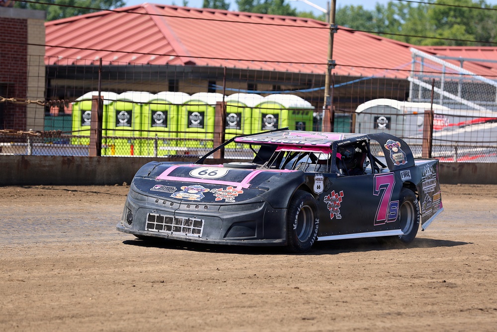 A black and pink dirt track racing car numbered '7s' is competing on a dirt oval track. The modified stock car features Route 66 branding on the hood and multiple sponsor decals on the body. The background shows portable toilets, fencing, and a building with a red roof typical of a small regional racing venue.