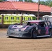 A black and pink dirt track racing car numbered '7s' is competing on a dirt oval track. The modified stock car features Route 66 branding on the hood and multiple sponsor decals on the body. The background shows portable toilets, fencing, and a building with a red roof typical of a small regional racing venue.