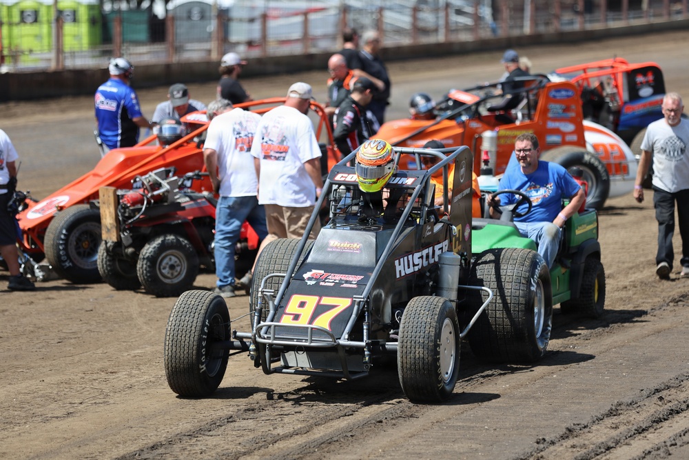 A dirt track racing event showing multiple sprint cars and midget race cars in a pit or staging area. The foreground features car number 97 with 'HUSTLER' branding being towed by a green utility vehicle, with the driver wearing a yellow/orange helmet. Several crew members, spectators, and orange sprint cars are visible in the background on a dirt surface.
