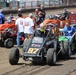 A dirt track racing event showing multiple sprint cars and midget race cars in a pit or staging area. The foreground features car number 97 with 'HUSTLER' branding being towed by a green utility vehicle, with the driver wearing a yellow/orange helmet. Several crew members, spectators, and orange sprint cars are visible in the background on a dirt surface.