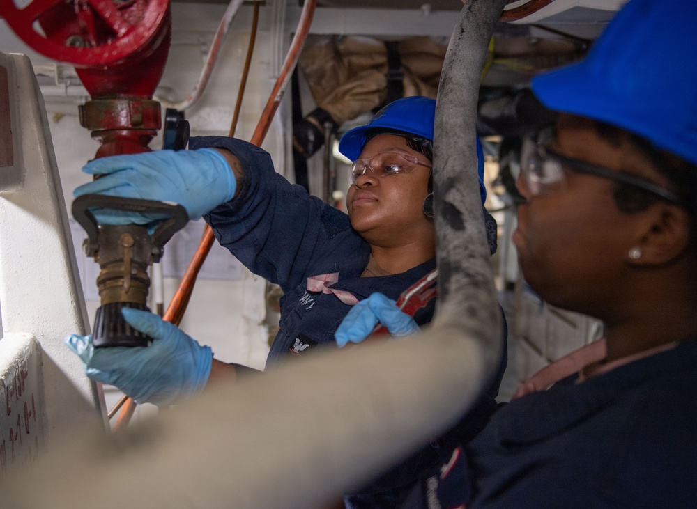 Two individuals wearing blue hard hats, safety glasses, and light blue protective gloves are working on industrial piping or valve equipment in a confined mechanical space. The person in the foreground is partially out of focus while the person in the background is actively manipulating a valve or fitting connected to red and copper-colored pipes. The setting appears to be aboard a naval vessel or industrial facility based on the white-painted metal walls and pipe infrastructure.