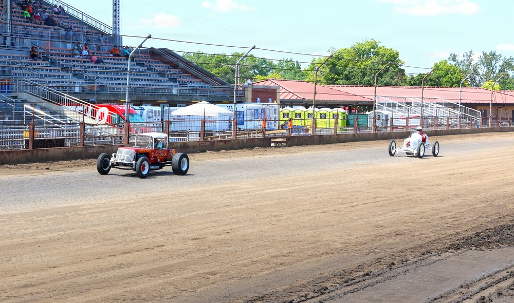 Two vintage racing cars are competing on a dirt oval track at what appears to be a county fair or small regional racing venue. One car is red and resembles a classic hot rod or T-bucket style vehicle, while the other is a white open-wheel midget or sprint-style racer. Spectators are visible in the grandstands on the left side, and various support structures and portable facilities are visible in the background.