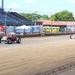 Two vintage racing cars are competing on a dirt oval track at what appears to be a county fair or small regional racing venue. One car is red and resembles a classic hot rod or T-bucket style vehicle, while the other is a white open-wheel midget or sprint-style racer. Spectators are visible in the grandstands on the left side, and various support structures and portable facilities are visible in the background.