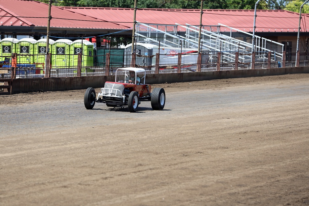 A vintage or modified open-wheel racing car is driving on a dirt oval track, likely during a pre-race preparation or practice session. The facility appears to be a small regional dirt track with grandstand seating, a red-roofed building, and portable toilets visible in the background. The track surface appears freshly groomed and the venue is not yet filled with spectators.