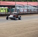A vintage or modified open-wheel racing car is driving on a dirt oval track, likely during a pre-race preparation or practice session. The facility appears to be a small regional dirt track with grandstand seating, a red-roofed building, and portable toilets visible in the background. The track surface appears freshly groomed and the venue is not yet filled with spectators.