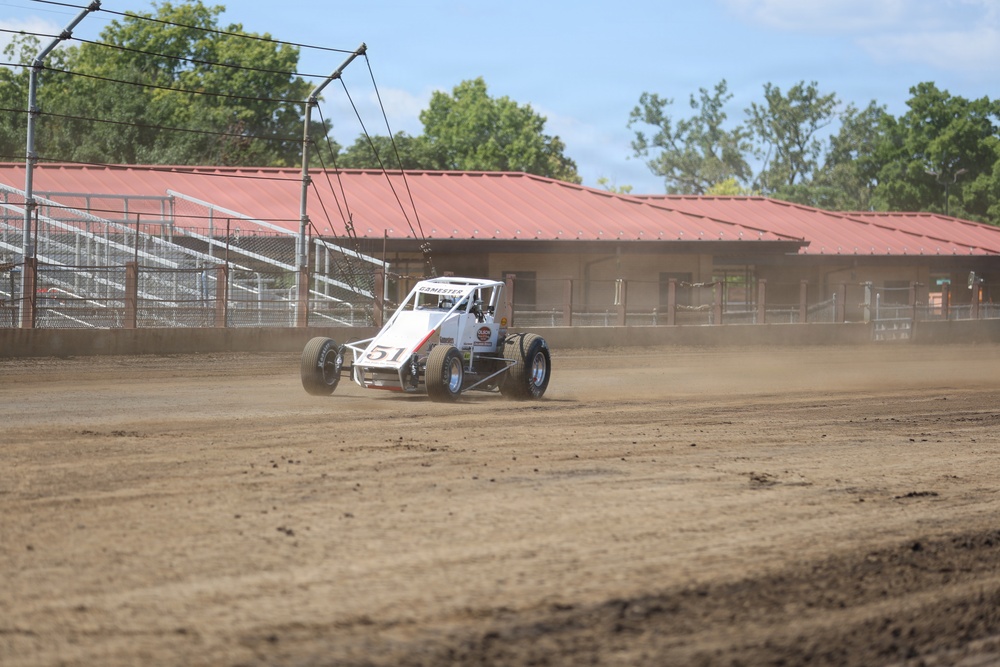 A white and red open-wheel dirt track racing car numbered 51 is navigating a dirt oval track, kicking up dust behind it. The track appears to be at a small regional racing facility with metal bleachers and a red-roofed building visible in the background. The grandstands are empty, suggesting this may be a practice session or pre-race activity.