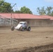 A white and red open-wheel dirt track racing car numbered 51 is navigating a dirt oval track, kicking up dust behind it. The track appears to be at a small regional racing facility with metal bleachers and a red-roofed building visible in the background. The grandstands are empty, suggesting this may be a practice session or pre-race activity.