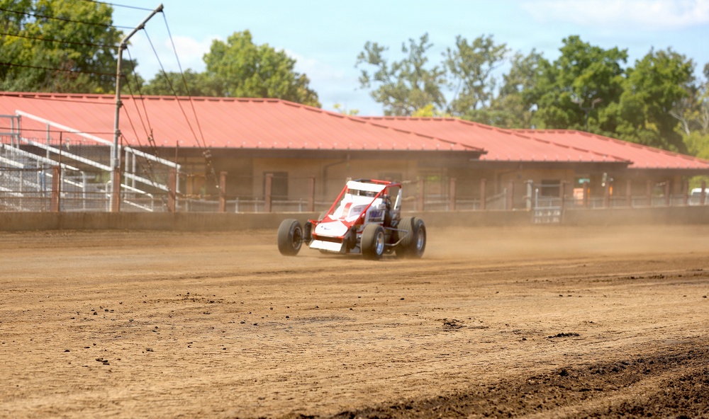 A red and white open-wheel dirt track racing car (wingless sprint car or midget racer) is navigating a dirt oval track, kicking up a dust cloud behind it. The setting appears to be a fairgrounds-style racing facility with a red-roofed building and metal bleachers/fencing visible in the background. Green trees line the perimeter under a partly cloudy sky.