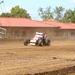 A red and white open-wheel dirt track racing car (wingless sprint car or midget racer) is navigating a dirt oval track, kicking up a dust cloud behind it. The setting appears to be a fairgrounds-style racing facility with a red-roofed building and metal bleachers/fencing visible in the background. Green trees line the perimeter under a partly cloudy sky.