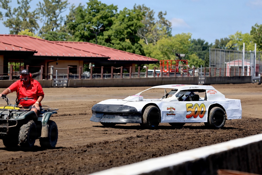 A white dirt track race car numbered '500' is on a dirt oval racing surface, accompanied by a man in a red shirt riding a green ATV. The setting appears to be a small local fairground or county fair racing venue, with a red-roofed building and trees visible in the background. The scene suggests pre-race preparation or a track inspection lap.