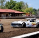 A white dirt track race car numbered '500' is on a dirt oval racing surface, accompanied by a man in a red shirt riding a green ATV. The setting appears to be a small local fairground or county fair racing venue, with a red-roofed building and trees visible in the background. The scene suggests pre-race preparation or a track inspection lap.