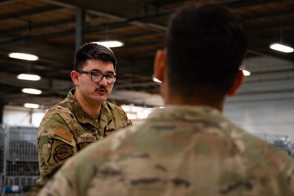 Two military personnel in OCP (Operational Camouflage Pattern) uniforms are engaged in conversation inside what appears to be a military hangar or warehouse facility. The soldier facing the camera, identified by a name tape reading 'THOMPSON,' is wearing glasses and has a small mustache. The setting suggests a briefing, training, or informal discussion in an indoor military work environment.