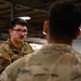 Two military personnel in OCP (Operational Camouflage Pattern) uniforms are engaged in conversation inside what appears to be a military hangar or warehouse facility. The soldier facing the camera, identified by a name tape reading 'THOMPSON,' is wearing glasses and has a small mustache. The setting suggests a briefing, training, or informal discussion in an indoor military work environment.