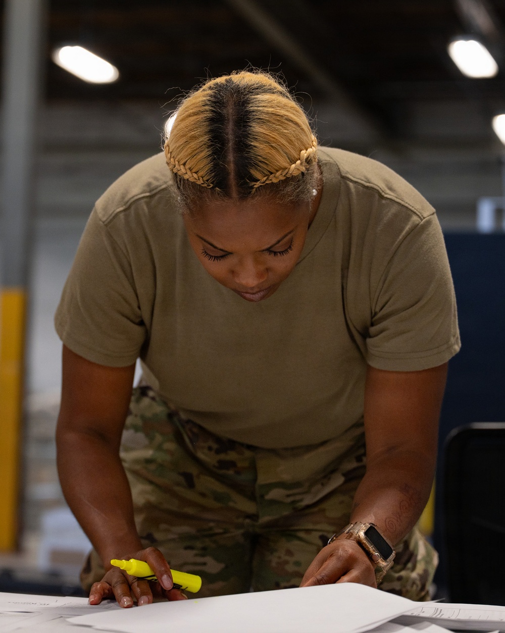 A female military service member in OCP (Operational Camouflage Pattern) uniform is leaning over a table, reviewing documents in what appears to be a warehouse or hangar facility. She is holding a yellow highlighter and appears to be actively marking or reviewing paperwork. The image is a close-up, professionally composed photograph with sharp focus on the subject.