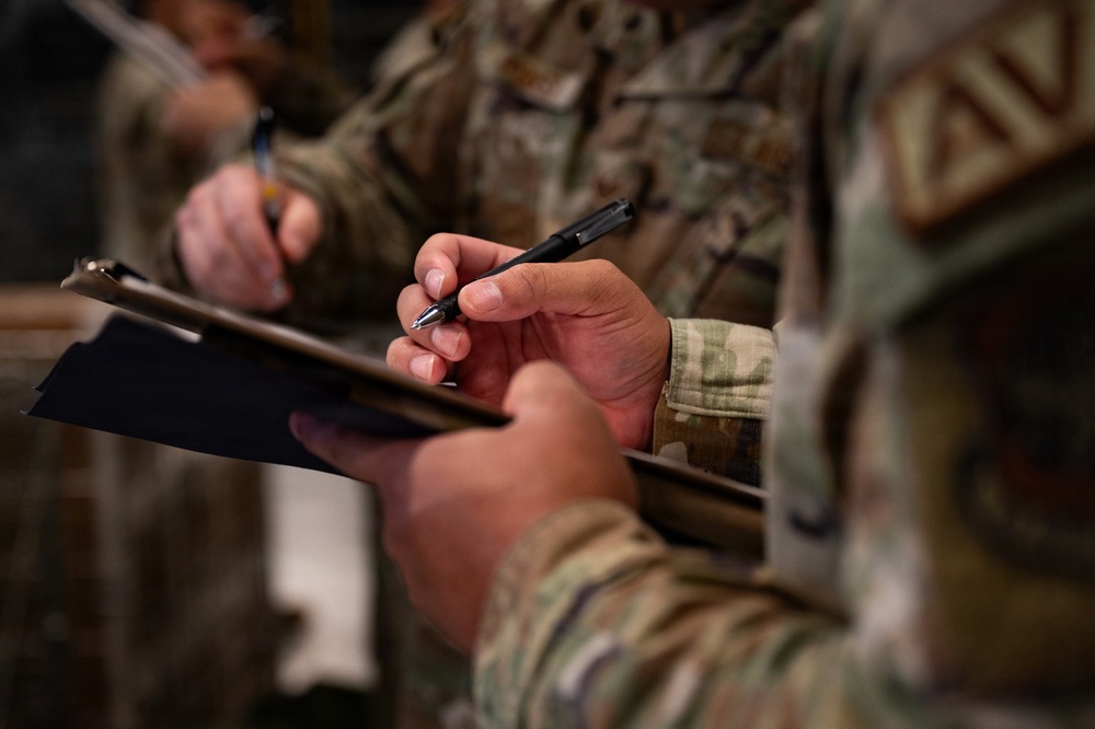 The image shows military personnel in camouflage uniforms writing on clipboards, likely during an administrative or operational task. At least two individuals are visible, both holding pens and clipboards simultaneously. The focus is on the foreground subject's hands, with the background figure slightly out of focus.