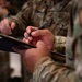 The image shows military personnel in camouflage uniforms writing on clipboards, likely during an administrative or operational task. At least two individuals are visible, both holding pens and clipboards simultaneously. The focus is on the foreground subject's hands, with the background figure slightly out of focus.