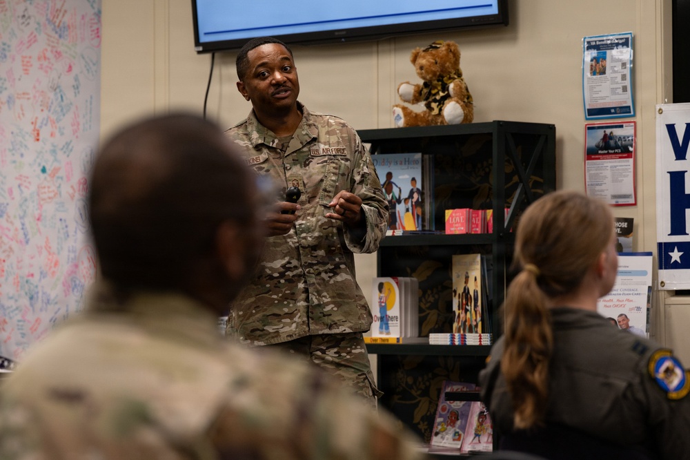 A U.S. Air Force service member in OCP (Operational Camouflage Pattern) uniform is addressing a small group of military personnel in what appears to be a resource or community support center. The speaker is standing and gesturing while presenting to seated audience members. The setting includes a bookshelf with books and a teddy bear, informational posters, and a wall covered in signatures or messages.