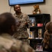 A U.S. Air Force service member in OCP (Operational Camouflage Pattern) uniform is addressing a small group of military personnel in what appears to be a resource or community support center. The speaker is standing and gesturing while presenting to seated audience members. The setting includes a bookshelf with books and a teddy bear, informational posters, and a wall covered in signatures or messages.